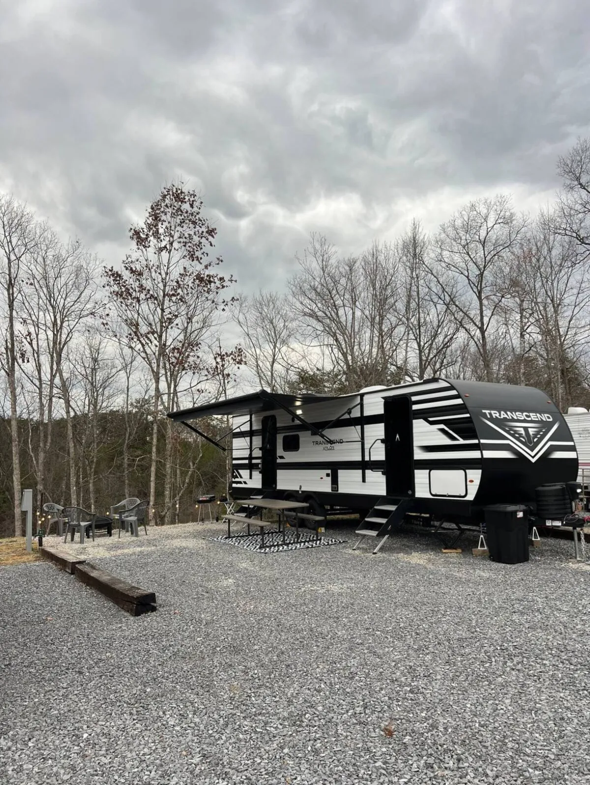 The image features a gleaming, modern travel trailer nestled amid tall, peaceful trees. A cozy picnic table and inviting outdoor seating are set up beside the trailer, suggesting space for meals, relaxation, and enjoying the natural surroundings. The lush greenery around the camper hints at privacy and immersion in nature, perfect for a serene getaway.