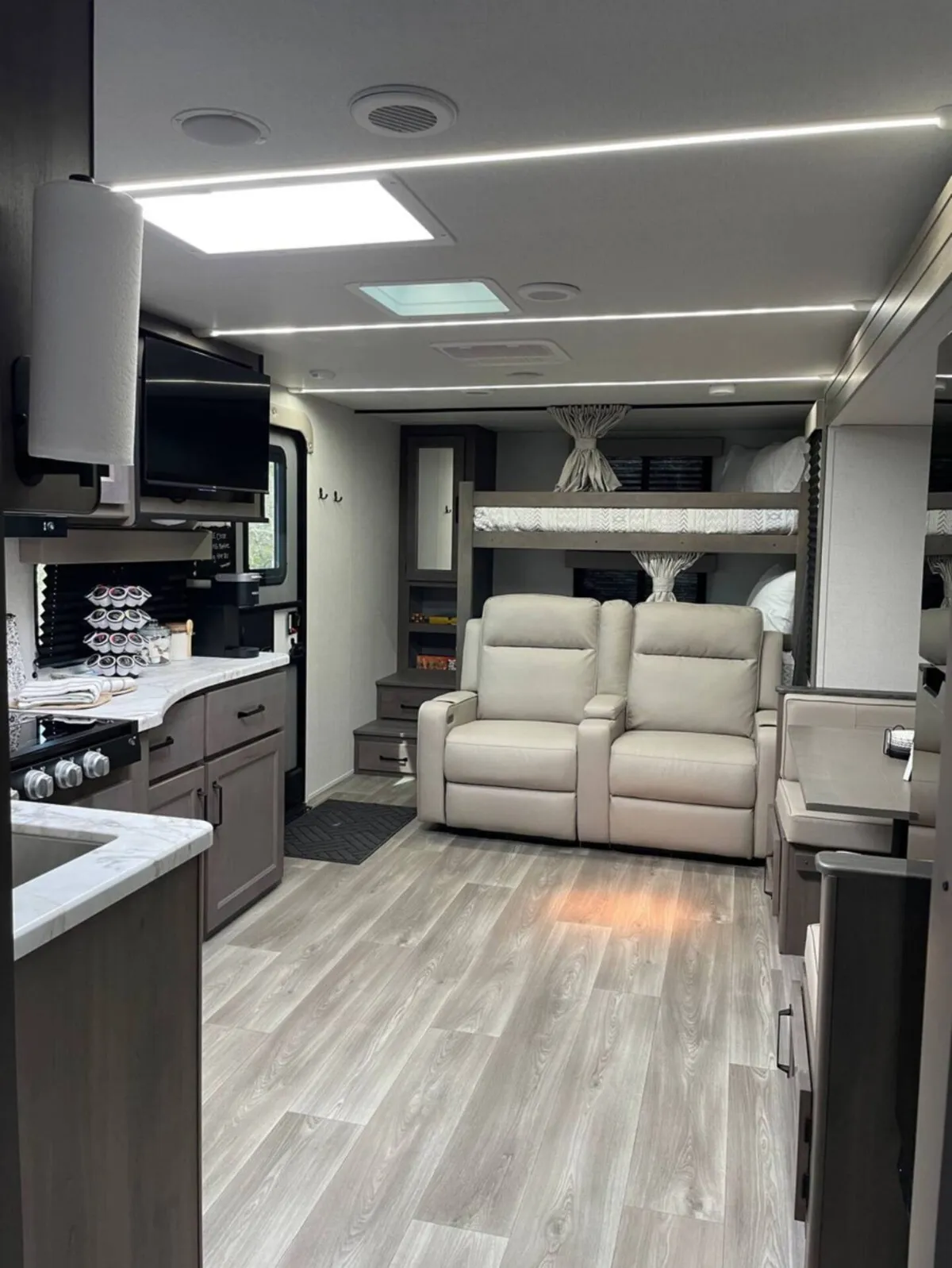 This image showcases the living area of the RV, with two reclining chairs positioned near a window, the adjacent kitchen area, and nearby bunk beds. The space is arranged to invite relaxation, conversation, and togetherness after a day of adventures.