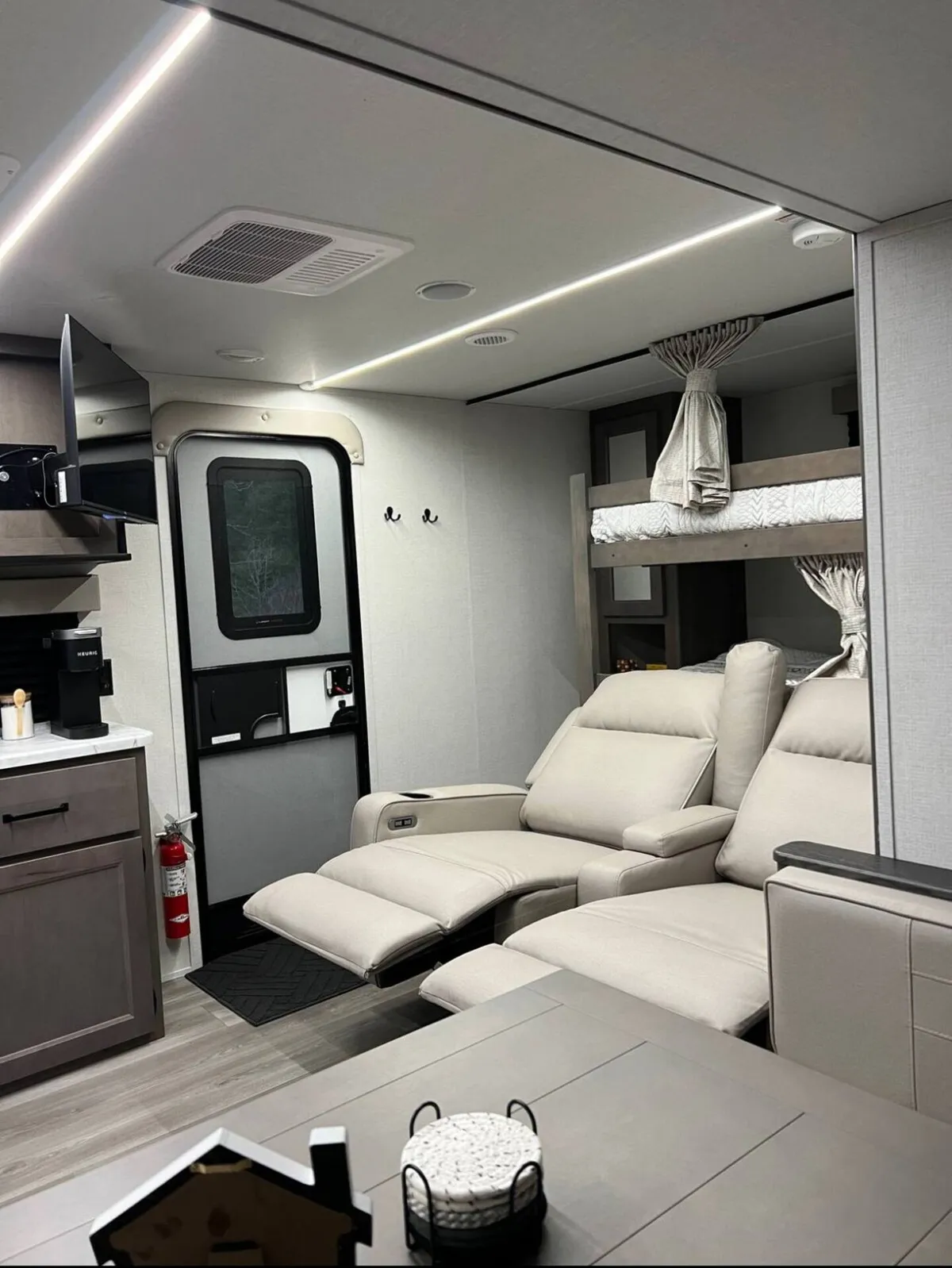 A broad view of the interior, featuring both the reclining chairs and the dinette alongside the kitchen and bunk area. The arrangement encourages gatherings around the dining table and relaxation in the recliners, all under modern, soft lighting.