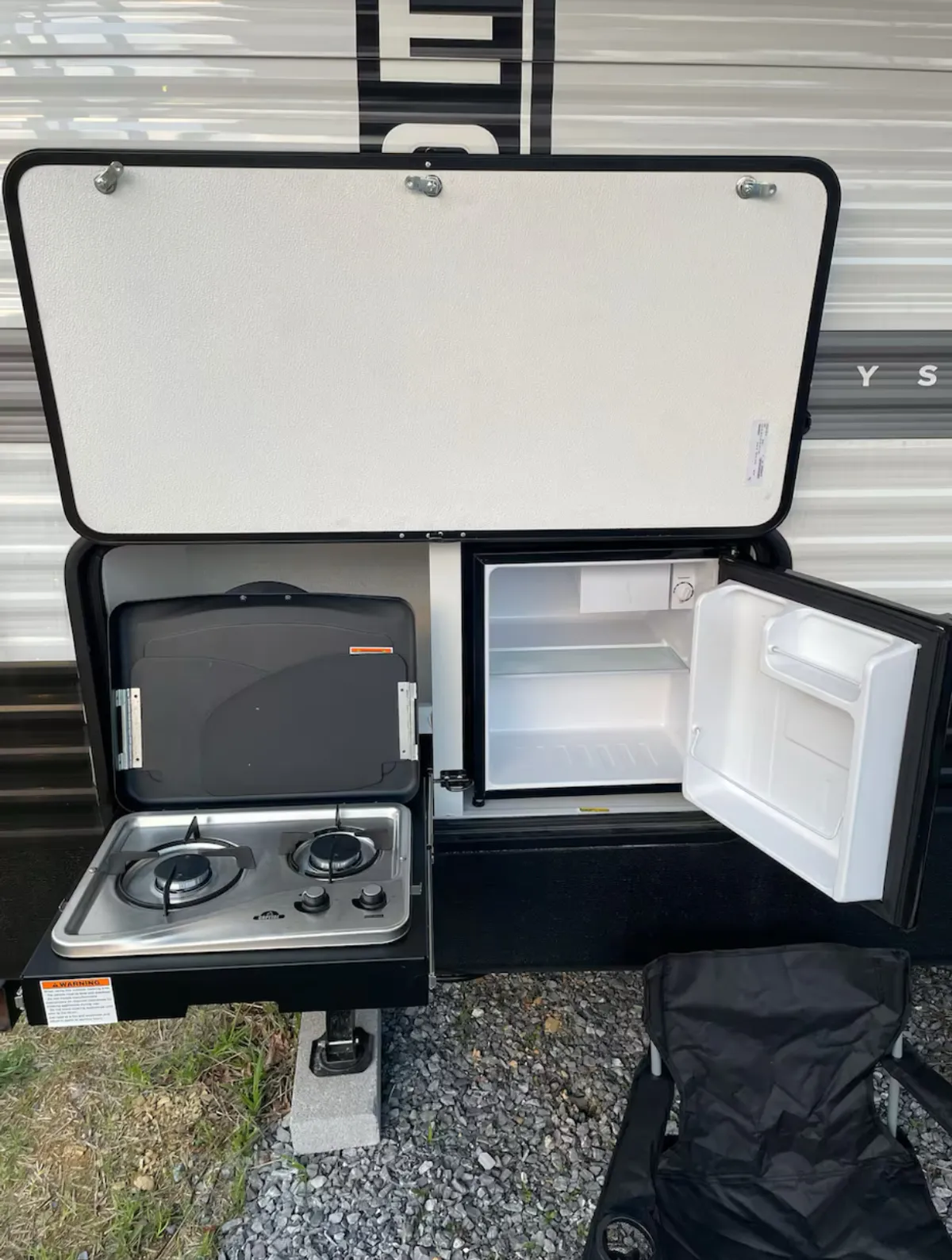 This image captures an inviting outdoor hangout area with a stove, mini refrigerator, and comfortable chair arranged under the open sky. The scene conveys a casual, adventurous mood, complete with setup for outdoor dining, shared laughter, and relaxation around a campfire.