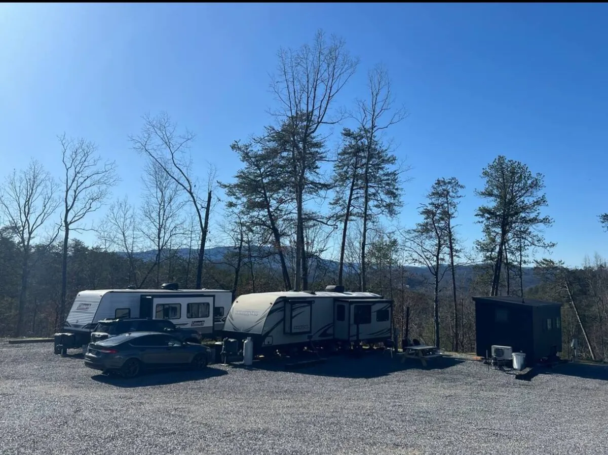 A scenic exterior shot showcasing two RVs nestled among trees with a car parked nearby. The mountain backdrop and relaxed outdoor setup evoke a sense of calm and adventure, perfect for morning coffee or stargazing after a day of outdoor activities.